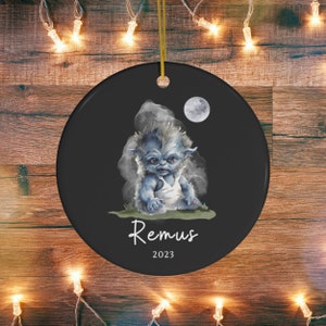 Pu&ograve; includere: Ornamento natalizio rotondo in ceramica nera con un'illustrazione di una creatura blu, il nome "Remus" e l'anno "2023". Il design include una luna piena e un cordino dorato per appenderlo. Ideale per le festivit&agrave;.