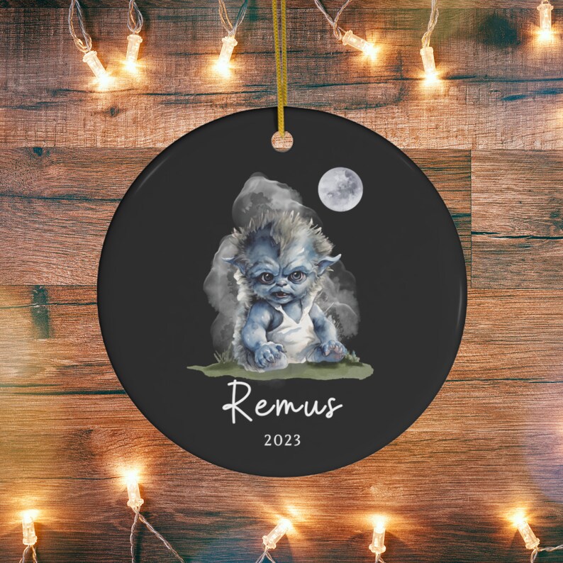 Pu&ograve; includere: Ornamento natalizio rotondo in ceramica nera con un'illustrazione di una creatura blu, il nome "Remus" e l'anno "2023". Il design include una luna piena e un cordino dorato per appenderlo. Ideale per le festivit&agrave;.