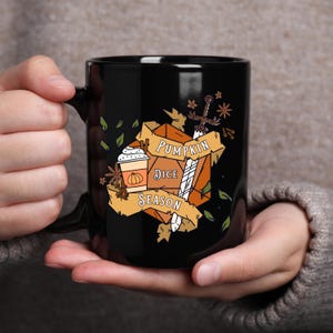 May include: Black ceramic mug featuring a fall-themed design. The design includes a pumpkin spice latte, dice, a sword, and the words "Pumpkin Dice Season" on a banner. The mug is held by a person.