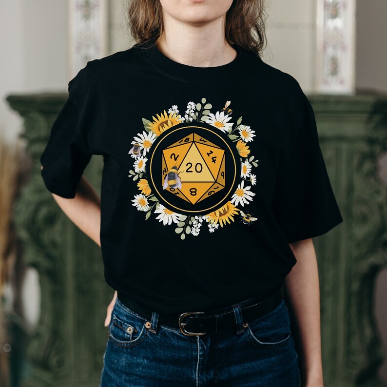 Może przedstawiać: Czarny T-shirt z ż&oacute;łtą 20-ścienną kostką do gry otoczoną białymi stokrotkami i słonecznikami. Na kostce siedzi pszczoła. Tekst na kostce brzmi "20, 12, 14, 16, 18, 9, 6, 8, 4, 2, 1, 3, 5, 7, 10, 19, 17, 15, 13, 11".