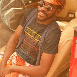 May include: A person wearing a black t-shirt with the text "Moony Formal Foot Rongs" in a retro rainbow design, sitting in a vintage car. The person is wearing a brown leather cap and round sunglasses.
