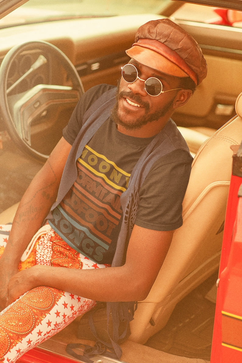 May include: A person wearing a black t-shirt with the text "Moony Formal Foot Rongs" in a retro rainbow design, sitting in a vintage car. The person is wearing a brown leather cap and round sunglasses.