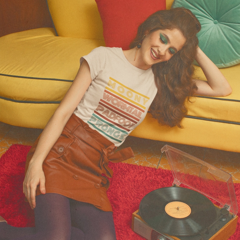 May include: A woman wearing a white t-shirt with the text "Moony Wormtail Padfoot Prongs" in colorful stripes. She is sitting on a yellow couch with a brown skirt and purple tights. She is listening to a record player with a black vinyl record.