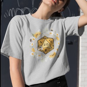 Może przedstawiać: Szary T-shirt z ż&oacute;łtą 20-ścienną kostką do gry otoczoną białymi stokrotkami i ż&oacute;łtymi słonecznikami. Na kostce siedzi pszczoła.