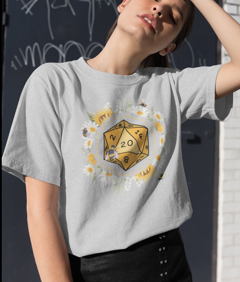 Może przedstawiać: Szary T-shirt z ż&oacute;łtą 20-ścienną kostką do gry otoczoną białymi stokrotkami i ż&oacute;łtymi słonecznikami. Na kostce siedzi pszczoła.