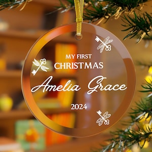 Op de afbeelding: Een heldere, ronde kerstbal met een gouden lint. De bal heeft de tekst "MY FIRST CHRISTMAS Amelia Grace 2024" in witte letters, samen met sleutelontwerpen van libellen.