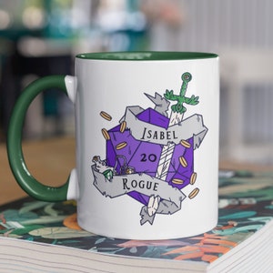 以下が含まれることがあります： 緑色の縁取りと取っ手のある白いセラミック製のマグカップ。マグカップには、剣、コイン、巻物のある紫色の20面ダイスが描かれています。巻物には「Isabel Rogue 20」と書いてあります。