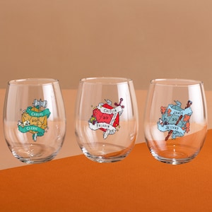 Może przedstawiać: Trzy bezszypułkowe kieliszki do wina z kolorową grafiką. Każdy kieliszek ma inny wzór z imieniem, kostką i tytułem klasy. Kieliszki mają przezroczystą, gładką powierzchnię i idealnie nadają się do napojów.