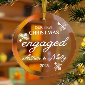 Op de afbeelding: Een heldere, ronde kerstversiering met gouden rand en een gouden lint. De versiering toont de tekst "OUR FIRST CHRISTMAS engaged Arthur & Molly 2023" in witte letters, met sleutel- en libellenontwerpen.