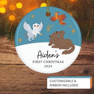 Op de afbeelding: Een ronde kerstversiering met een blauwe achtergrond met cartoon illustraties van een vogel, een feniks en een driekoppige hond. De tekst "Aiden's FIRST CHRISTMAS 2024" is weergegeven. Gouden lint om op te hangen.
