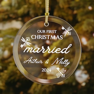 Op de afbeelding: Een heldere, ronde kerstbal met gouden rand en een gouden lint. De bal heeft de tekst "OUR FIRST CHRISTMAS married Arthur & Molly 2024" in witte letters, samen met libellen- en sleutelontwerpen.