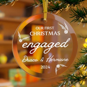 Op de afbeelding: Een heldere, ronde kerstversiering met gouden rand en een gouden lint. De versiering leest "OUR FIRST CHRISTMAS engaged Draco & Hermione 2024" in witte letters. Decoratieve vleugels zijn ook afgebeeld.