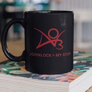 K&ouml;nnte beinhalten: Schwarze Keramik-Kaffeetasse mit einer roten Grafik einer Person mit dem Text "JOHNLOCK = MY OTP" auf der Vorderseite.
