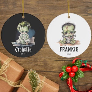 Pu&ograve; includere: Due ornamenti natalizi rotondi. Uno &egrave; nero con un bambino dalla pelle verde di nome "Ophelia" e l'anno "2023". L'altro &egrave; bianco con un bambino dalla pelle verde di nome "Frankie" e l'anno "2023". Entrambi hanno ganci dorati.