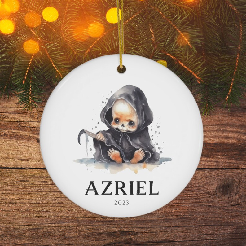 Pu&ograve; includere: Ornamento natalizio rotondo in ceramica bianca con un'illustrazione ad acquerello di un bambino mietitore con un mantello nero con cappuccio, che tiene una falce. Il nome "AZRIEL" e l'anno "2023" sono stampati sotto. &Egrave; attaccato un filo d'oro.