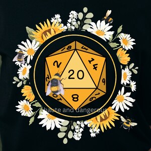 Może przedstawiać: Czarny T-shirt z ż&oacute;łtą 20-ścienną kostką do gry otoczoną ż&oacute;łtymi i białymi kwiatami i pszczołami. Tekst "haute and dangerous" jest nadrukowany pod kostką.