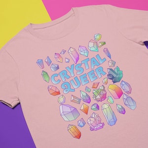 Puede incluir: Una camiseta rosa con el texto "CRYSTAL QUEER" en letras azules. El texto está rodeado de una variedad de cristales coloridos.
