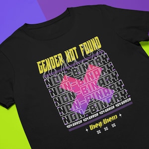 May include: Black t-shirt featuring a graphic design that reads "Gender Not Found" with the words "Non-Binary" repeated multiple times in a stacked pattern. The graphic also includes a pink and purple X with flames. The text "404 Error 404 Error 404 Error 404 Error" is printed below the graphic. The text "they/them" is printed below the error message.