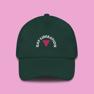 Op de afbeelding: Een donkergroene baseballpet met een wit geborduurd ontwerp dat "Gay Liberation" zegt met een roze driehoek in het midden.