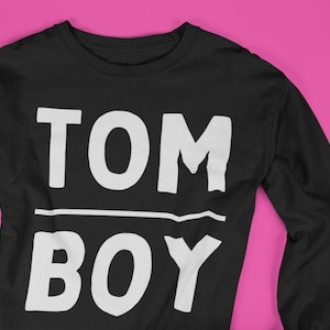 May include: Black long-sleeve shirt with white text that reads "TOMBOY" in a bold, blocky font.