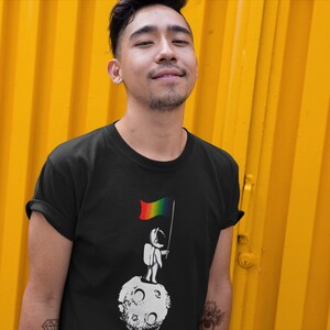 May include: A person wearing a black t-shirt with a graphic of an astronaut standing on the moon holding a rainbow flag.