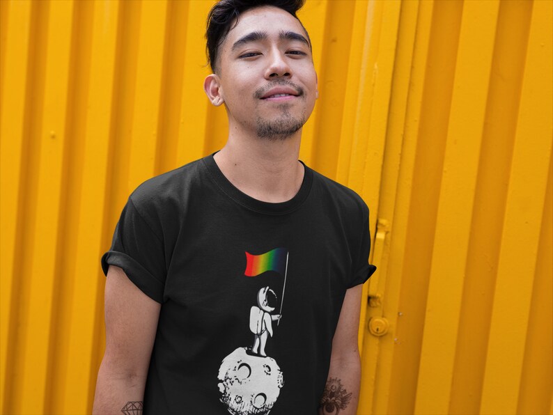 May include: A person wearing a black t-shirt with a graphic of an astronaut standing on the moon holding a rainbow flag.