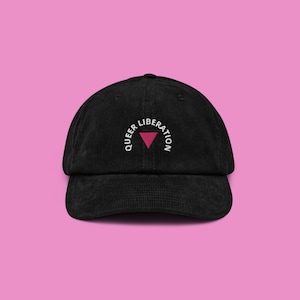 Peut inclure: Casquette de baseball en velours côtelé noir avec l'inscription "QUEER LIBERATION" brodée en blanc, arquée au-dessus d'un triangle rose. Le chapeau est posé sur un fond rose.