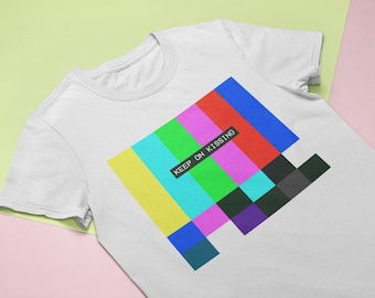 May include: White t-shirt featuring a colourful pixelated design and the text "KEEP ON KISSING" in black.