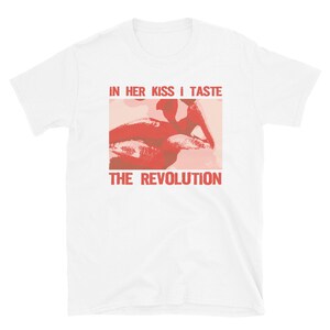 Pu&ograve; includere: Maglietta bianca con una grafica rossa e rosa di un paio di labbra. Il testo "IN HER KISS I TASTE THE REVOLUTION" &egrave; stampato in rosso sulla maglietta.