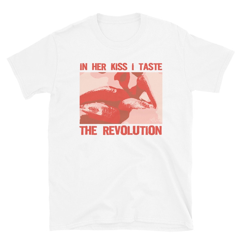 Pu&ograve; includere: Maglietta bianca con una grafica rossa e rosa di un paio di labbra. Il testo "IN HER KISS I TASTE THE REVOLUTION" &egrave; stampato in rosso sulla maglietta.