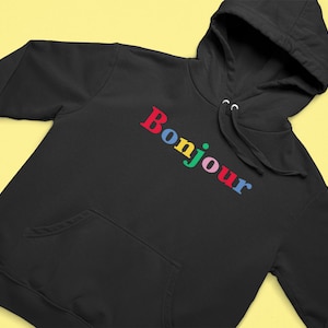 Puede incluir: Sudadera con capucha negra con la palabra "Bonjour" en colores arcoíris.