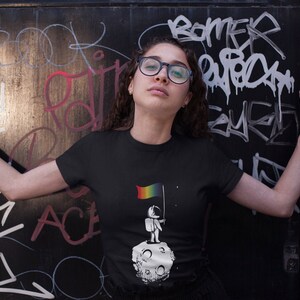 May include: A black t-shirt featuring a graphic of a person standing on the moon holding a rainbow flag. The text "One small step for humankind" is printed on the shirt.