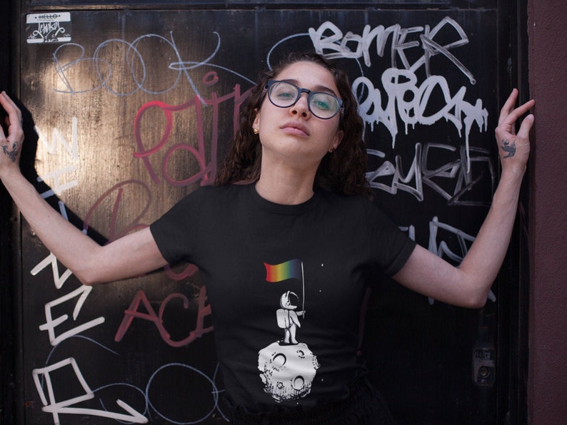 May include: A black t-shirt featuring a graphic of a person standing on the moon holding a rainbow flag. The text "One small step for humankind" is printed on the shirt.
