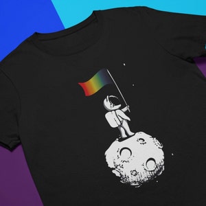 May include: A black t-shirt featuring a graphic of an astronaut standing on the moon holding a rainbow flag.
