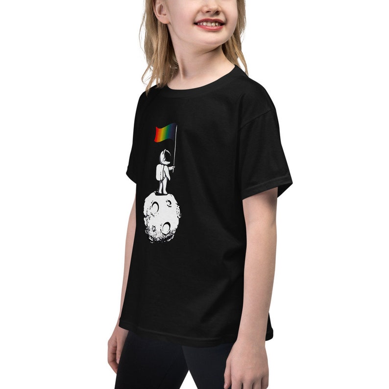 May include: A black youth t-shirt featuring a graphic of an astronaut standing on the moon holding a rainbow flag.