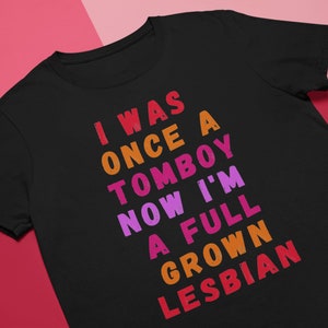 Puede incluir: Camiseta negra con un gráfico de arcoíris que dice "Yo era una vez una marimacho ahora soy una lesbiana adulta".