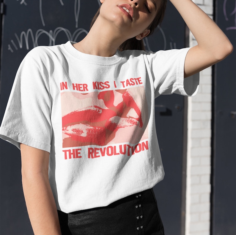 Pu&ograve; includere: Maglietta bianca con una grafica rossa e rosa di labbra e il testo "In her kiss I taste the revolution."