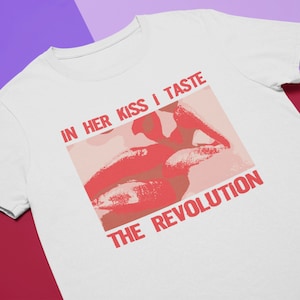 Pu&ograve; includere: Maglietta bianca con una grafica rossa di un paio di labbra e il testo "IN HER KISS I TASTE THE REVOLUTION".