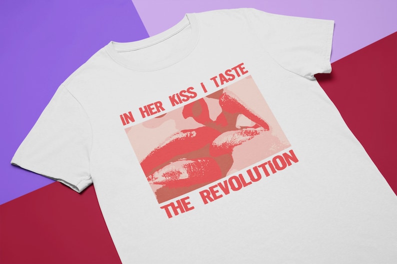 Pu&ograve; includere: Maglietta bianca con una grafica rossa di un paio di labbra e il testo "IN HER KISS I TASTE THE REVOLUTION".
