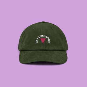 Peut inclure: Une casquette de baseball en velours côtelé vert avec le texte "Gay Liberation" brodé en blanc avec un symbole de triangle rose.