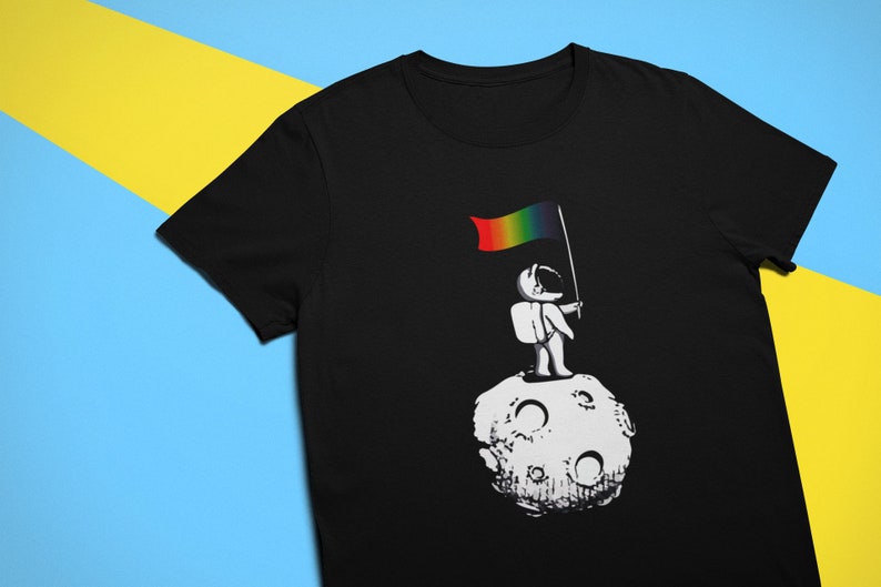May include: A black t-shirt featuring a graphic of an astronaut standing on the moon holding a rainbow flag.