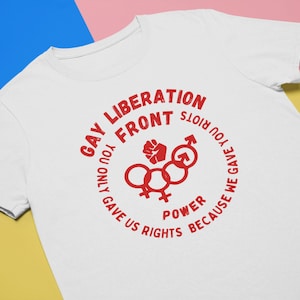 Può includere: T-shirt bianco con grafica rossa. La grafica include il testo "GAY LIBERATION FRONT" in un cerchio, insieme a simboli e al testo "YOU ONLY GAVE US RIGHTS BECAUSE WE GAVE YOU RIOTS".