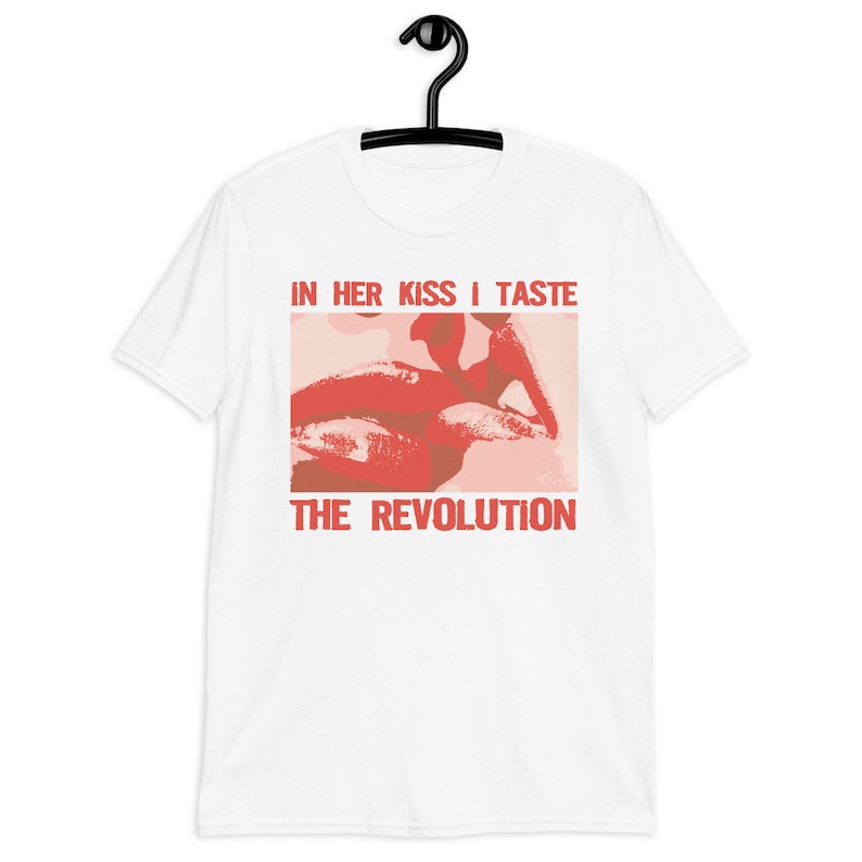 Pu&ograve; includere: Maglietta bianca con una grafica rossa e rosa di un primo piano di labbra con il testo "IN HER KISS I TASTE THE REVOLUTION"