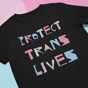 Può includere: Maglietta nera con il testo "PROTECT TRANS LIVES" in rosa, blu e bianco.