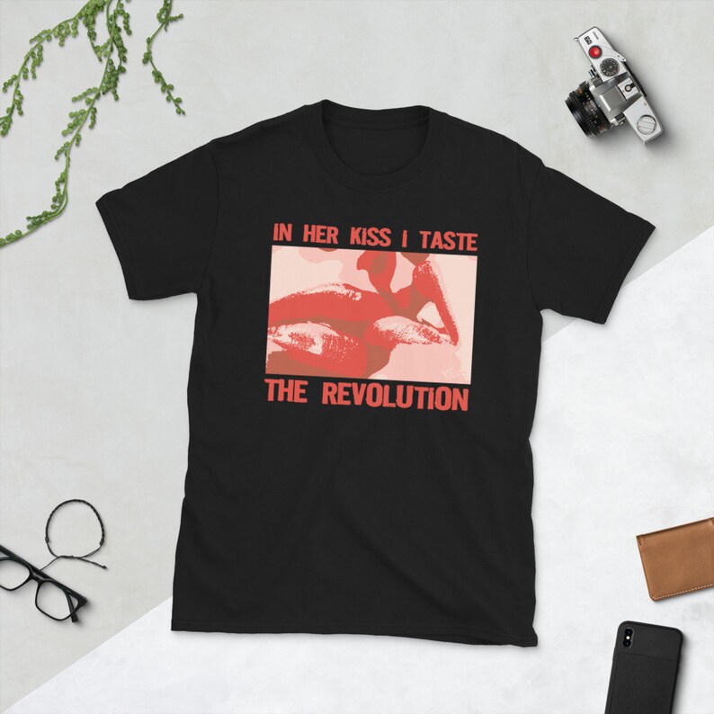 Pu&ograve; includere: Maglietta nera con una grafica rossa e rosa di una bocca con il testo "IN HER KISS I TASTE THE REVOLUTION" stampato su di essa.