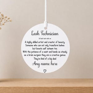 May include: A white, circular decoration featuring the words "Lash Technician" and a definition. The design includes space for a name and a heart symbol. It is suspended by a white string, set on a wooden surface.