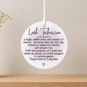 May include: A circular white ornament with the words "Lash Technician" in black script. The text includes a definition and description of a lash technician. The ornament is suspended by a white string.