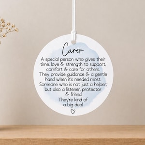 May include: A circular white ornament with the word "Carer" and a definition printed in black. It features a light blue watercolour design and a white string. A small heart is at the bottom of the text.