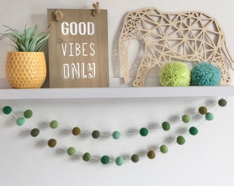 Può includere: Uno scaffale bianco con un cartello di legno che dice "Good Vibes Only" e una scultura di elefante in legno geometrico. Ci sono due pompon verdi e una pianta grassa in vaso sullo scaffale. Una ghirlanda di palline di feltro verde pende dallo scaffale.