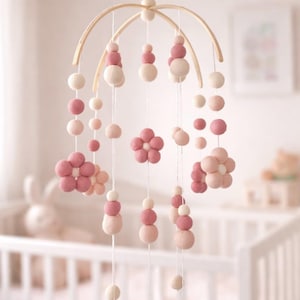 May include: A baby mobile with a wooden frame, featuring hanging pink and white felt balls and flower shapes. The mobile is suspended above a white crib in a soft, light-filled room.